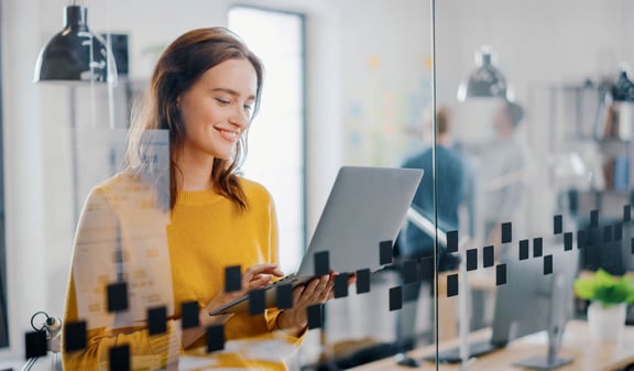 : Junge Frau steht lächelnd mit Laptop in der Hand hinter einer Glaswand – Symbolbild für ein modernes Recruitingmanagement